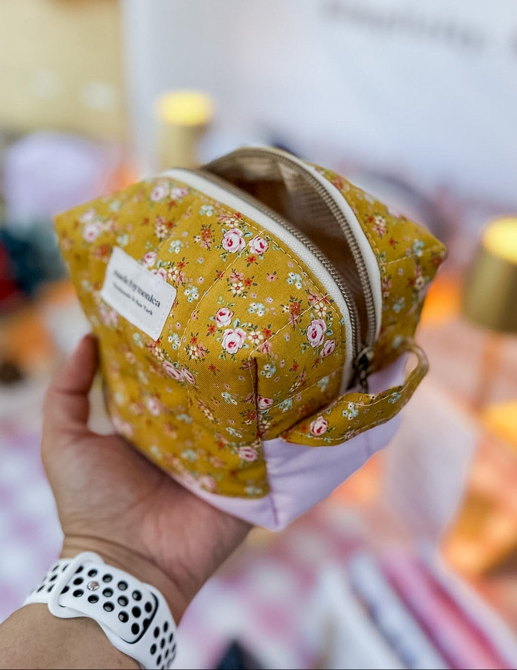 Hand holding a yellow fabric pouch with floral pattern in front of a blurred 'madeby' sign.